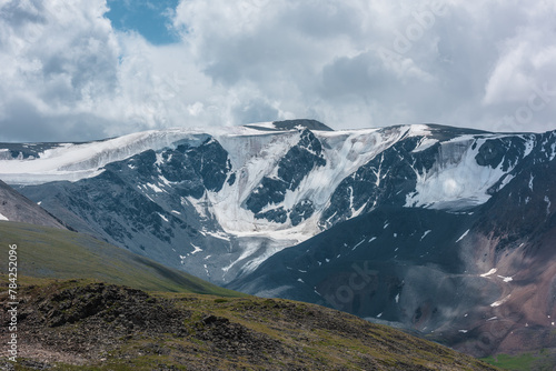 Fototapeta Naklejka Na Ścianę i Meble -  Dramatic view to large snow-capped mountain range in sunlight under gray cloudy sky. Atmospheric landscape with glacier on snowy rocky mountain wall. Shadows of clouds on rocks and high snow mountains