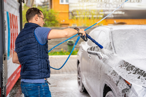 Young man washes his car at a self-service car wash using a hose with pressurized water and foam.