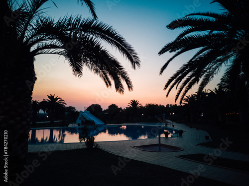 Soleil couchant sur une grande piscine avec les silhouettes des palmiers. Crépuscule sur un camping de vacances. Ambiance détente. Se reposer et admirer le paysage de la piscine et des palmiers. 
