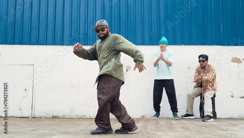 Asian hipster doing somersault in front of wall with lively friend. Attractive street dancer practicing break dance at street with blue background surrounded by friends. Outdoor sport 2024. Endeavor.