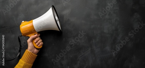 A hand in a mustard yellow sweater holding a megaphone against a textured black background, signaling a call to attention or announcement. photo