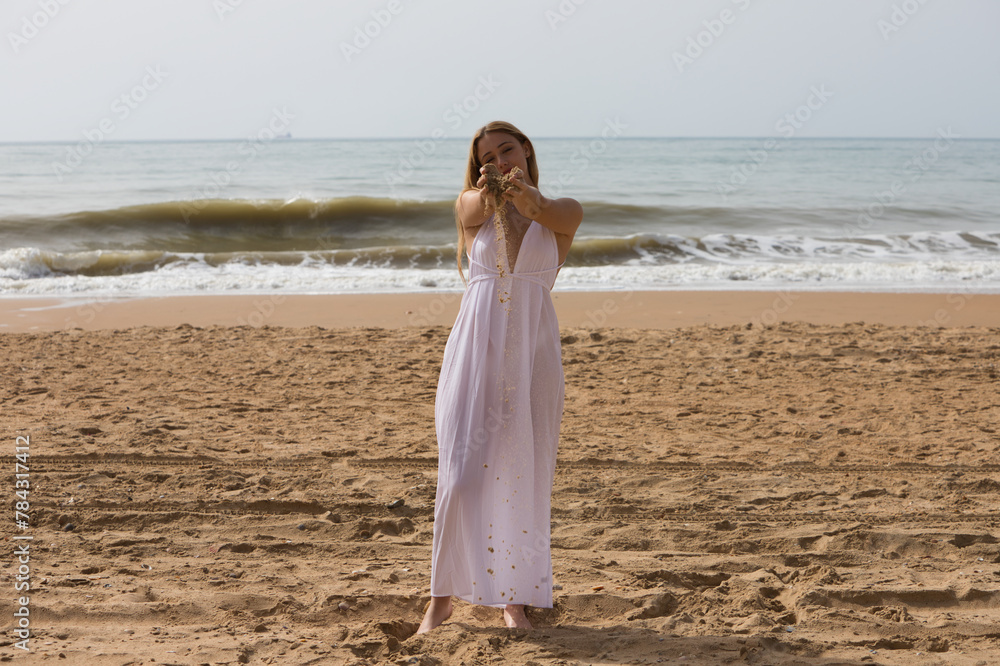 Pretty young blonde woman in white dress spills sand from between her hands as she holds her hands out in front of her. The woman is happy and having fun. In the background the blue sea. Pretty young blonde woman in white dress spills sand from between her hands as she holds her hands out in front of her. The woman is happy and having fun. In the background the blue sea.