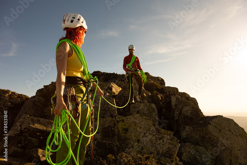 Two people are standing on a rocky mountain, one of them wearing a yellow shirt. They are holding green ropes and appear to be preparing for a climb