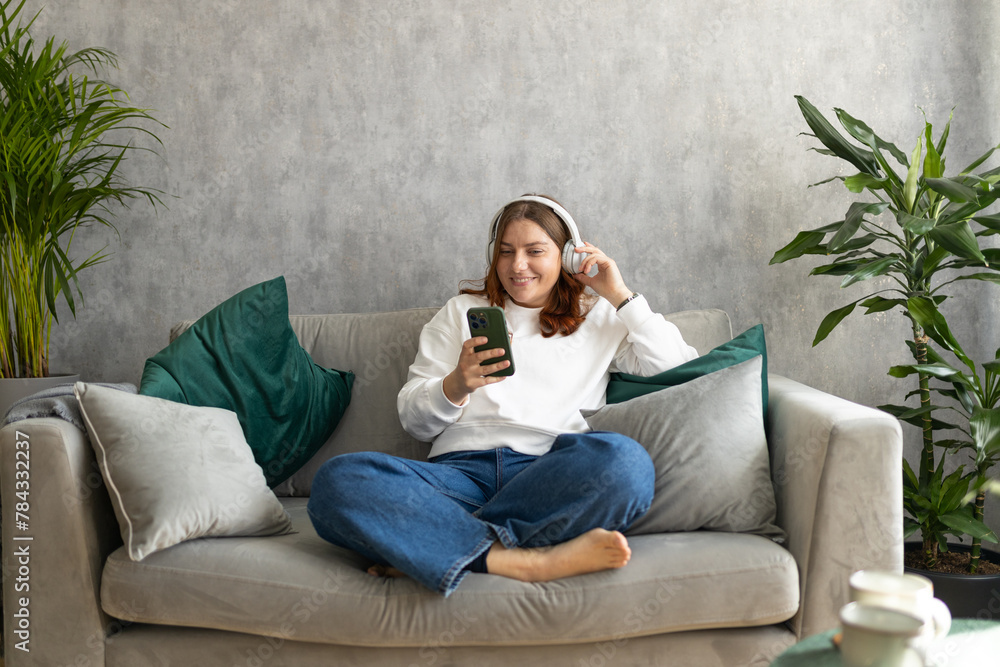 Smiling girl relaxing at home, she is listening to music, using a smartphone and wearing white headphones. Young 30s woman hearing enjoying good audio sound lounge on couch at home, full body