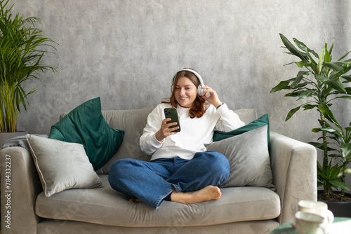 Obraz na płótnie Smiling girl relaxing at home, she is listening to music, using a smartphone and wearing white headphones. Young 30s woman hearing enjoying good audio sound lounge on couch at home, full body w salonie