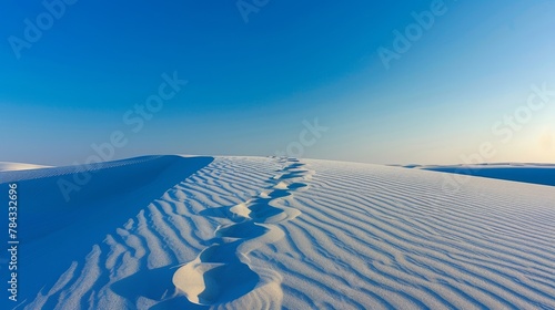 The image captures footprints meandering through a pristine sand dune under a clear blue sky, accentuating nature's beauty and the concept of journey photo