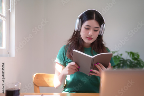 Young asian student women wearing headphone to studying and watching lecture webinar in online class on laptop while reading a book and learning knowledge remote education from home in living room