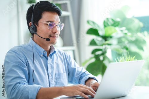 A man wearing a headset and glasses is typing on a laptop. He is focused on his work and he is in a professional setting