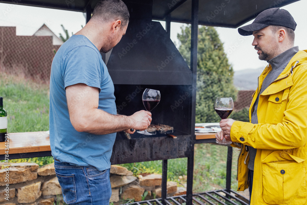 Two young men in casual clothes are roasting meat and drinking wine from glasses in backyard of house. Two young men in casual clothes are roasting meat and drinking wine from glasses in backyard of house.