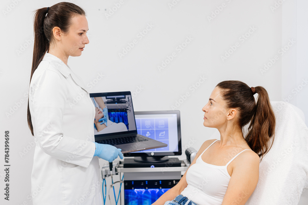 A female cosmetologist in a modern beauty salon holds a laptop with a broadcast of the hydrolifting procedure for facial cleansing. Cosmetic services advertising concept. A female cosmetologist in a modern beauty salon holds a laptop with a broadcast of the hydrolifting procedure for facial cleansing. Cosmetic services advertising concept.