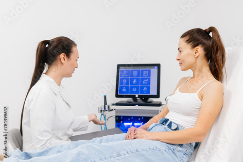 A female cosmetologist-dermatologist talks to a client sitting in a comfortable salon chair. Consultant cosmetologist advises on the facial hydrolifting procedure. Professional inspection.