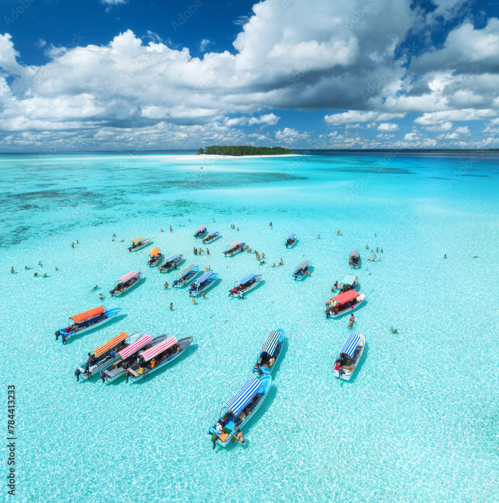 Aerial view of colorful boats in transparent water on sunny day. Mnemba island, Zanzibar. Top view of sandbank in low tide, blue sea, sand, swimming people, yachts, sky with clouds in summer. Ocean