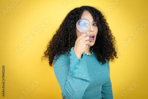 African american woman wearing casual sweater over yellow background surprised looking through a magnifying glass