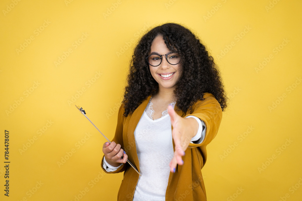 African american business woman with paperwork in hands over yellow background smiling friendly offering handshake as greeting and welcoming
