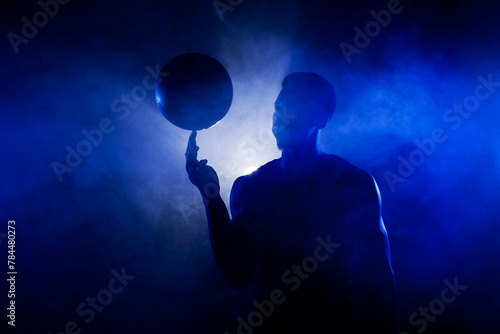 Basketball player silhouette lit with blue color spinning a ball against smoke background. Muscular african american man.
