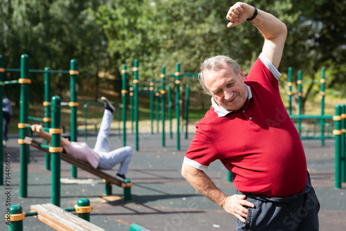 Happy joyful retired sportsman in sportswear doing exercises with arm above head, exercising outdoors in city park © caftor