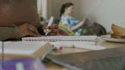 Cropped shot of unrecognizable African American female uni student highlighting notes in notebook while studying, co-living with Caucasian female roommate sitting on couch in blurred background