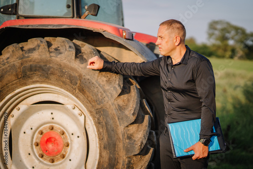 Fotografie Valmiera, Latvia - August 17, 2024 - A man stands beside a tractor tire in a field, holding a clipboard and examining the tire
