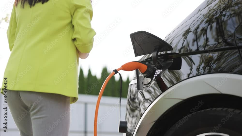 Woman connects an electric car to the charger and adjusts the process of charging the car battery using a cell phone smart phone. Girl plugs power cable to charge electric car in parking lot Woman connects an electric car to the charger and adjusts the process of charging the car battery using a cell phone smart phone. Girl plugs power cable to charge electric car in parking lot