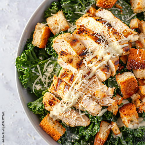 A close-up top-down view of a kale Caesar salad with grilled chicken, featuring crisp kale leaves tossed in creamy Caesar dressing, topped with grilled chicken strips, Parmesan cheese shavings, and cr