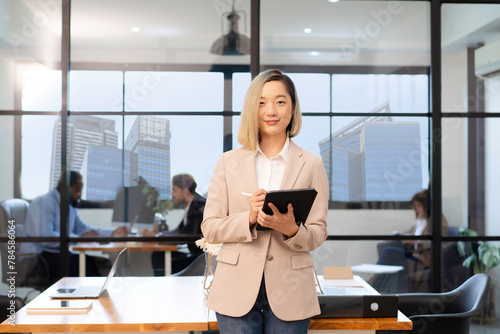 young asian businesswoman checklist with tablet in office with background of skyscrape view city building through window, group of multiracial team colleague diverse employee coworker meeting business