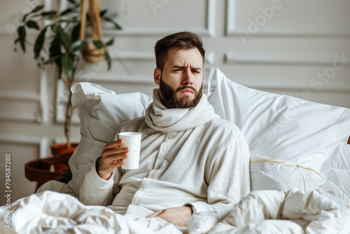 A young Caucasian man with a dark beard sick with a cold, flu, lying in a white bed at home, drinking medicine.