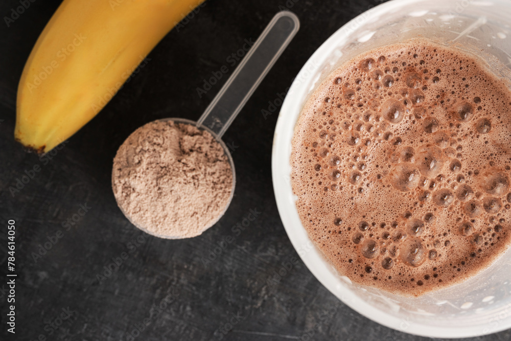 Blended chocolate protein drink in a shaker, plastic measuring spoon with protein powder, bananas on a dark background, top view Blended chocolate protein drink in a shaker, plastic measuring spoon with protein powder, bananas on a dark background, top view