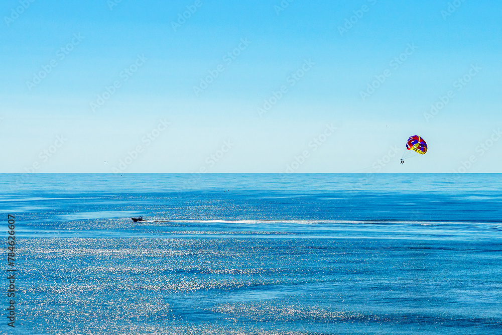 A vast expanse of the ocean gleams under the bright sky with a single Parasailing floating high above the sea, tethered to a distant boat. Myrtle Beach South Carolina US.