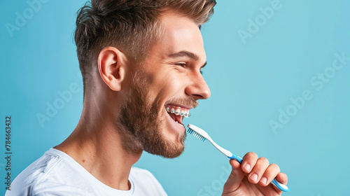 Young man in braces brushes his teeth, blue background copy space. Orthodontic device care concept, dental health, oral hygiene.