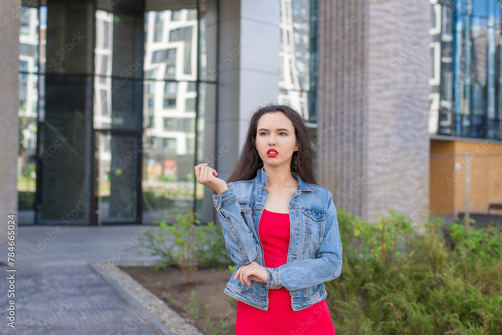 A young girl in a red dress stands in front of a building. She's wearing a denim jacket and red lipstick. A young girl in a red dress stands in front of a building. She's wearing a denim jacket and red lipstick.