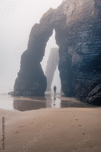 Man standing under natural arch on Cathedrals beach in Galicia, Spainn. Tourist silhouette in foggy landscape with Playa de Las Catedrales Catedrais beach in Ribadeo, Lugo on Cantabrian coast photo