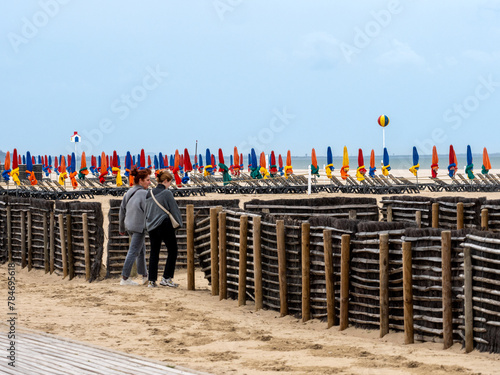 Deauville, France, April 5, 2023. Main promenade in Deauville on the beach, two women walking with their backs to the camera, in the background colorful beach umbrellas, blue red yellow