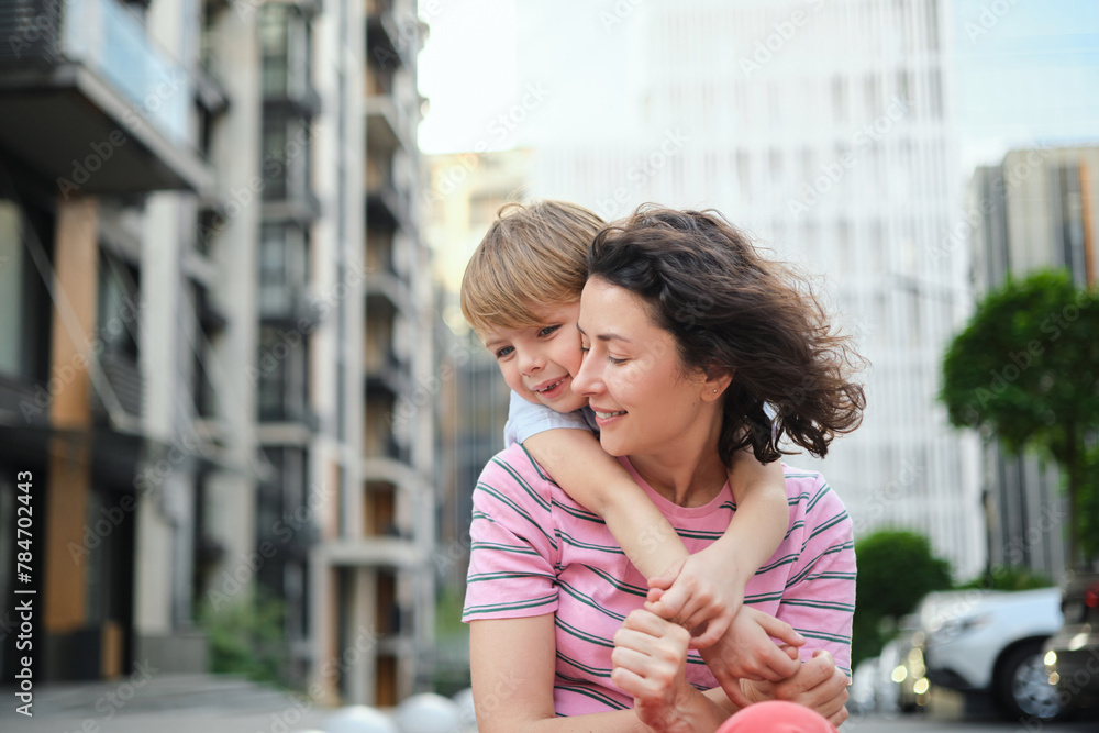 Fototapeta premium young mother and son smiling looking at the camera in modern residential complex. Having fun together, happy parenting, new home concept. Bright summer day in the urban background. High quality photo