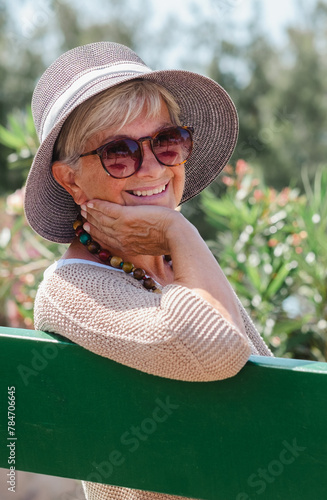 Portrait of beautiful senior woman wearing cap and glasses sitting outdoor on a bench of public park looking at camera. Smiling elderly female enjoying free moment in sunny day #784706645