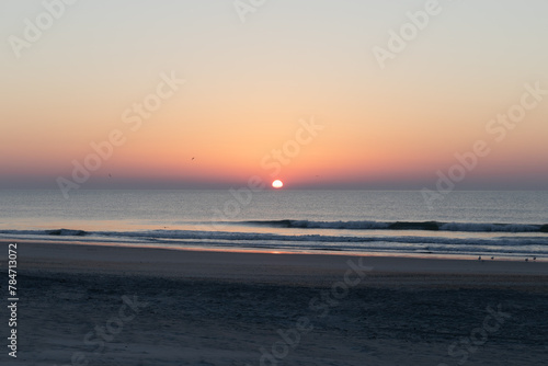 Colorful morning sun ascending over the ocean, deserted beach with calm waters, horizontal aspect