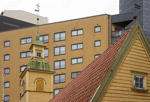 wooden house with a tiled roof and a turret with a weather vane against the background of modern buildings i