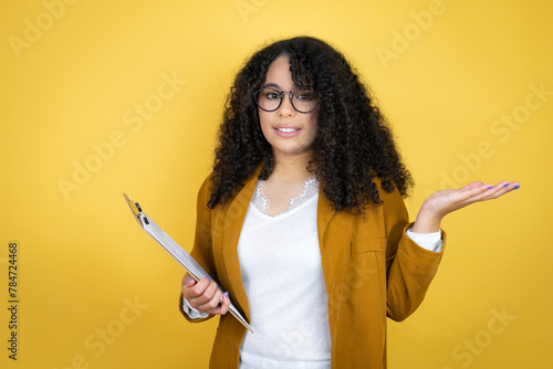 African american business woman with paperwork in hands over yellow background clueless and confused expression with arms and hands raised