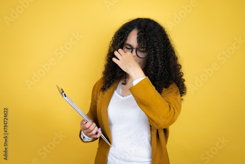 African american business woman with paperwork in hands over yellow background smelling something stinky and disgusting, intolerable smell, holding breath with fingers on nose photo