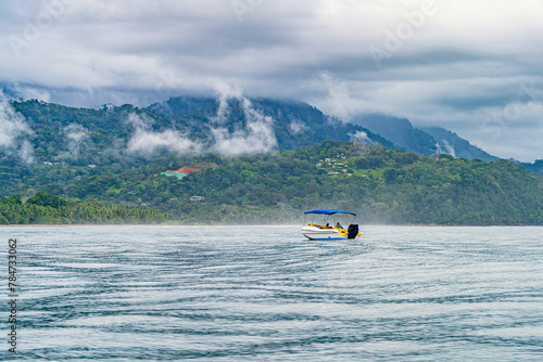 Leisure boat cruising on a serene sea with a misty mountainous backdrop and lush tropical forests. Tranquil ocean waters before a tropical coastline with clouds descending upon verdant hills. High