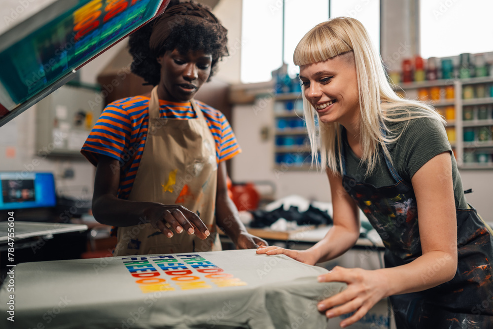 Fototapeta premium Happy interracial female print shop workers silkscreen printing on machine