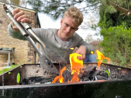 Selective focus on fire flames on bbq grill, happy man shuffling charcoal in the grill using tongs on background