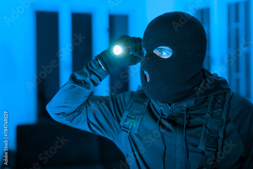 Masked burglar looking around in dark apartment photo