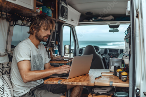 Man in summer clothes, sitting in a motorhome, parked in front of the beach, while working on his laptop