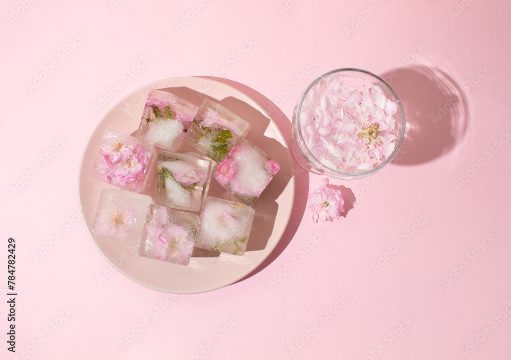 Beautiful small pink flowers in ice cubes and glass with petals on pink plate on pink background, top view Beautiful small pink flowers in ice cubes and glass with petals on pink plate on pink background, top view
