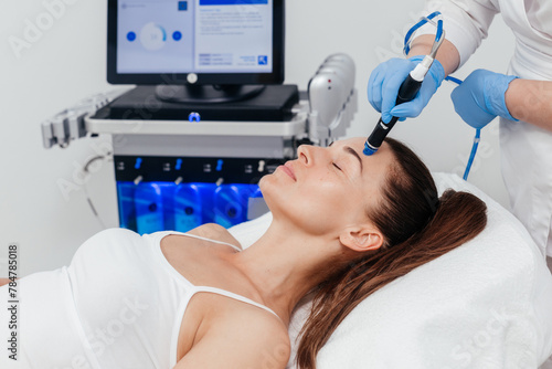 Middle aged beautician holding ultrasound device for hydro facial lifting and skin tightening procedure. Woman cosmetologist using modern cosmetology equipment in clinic photo