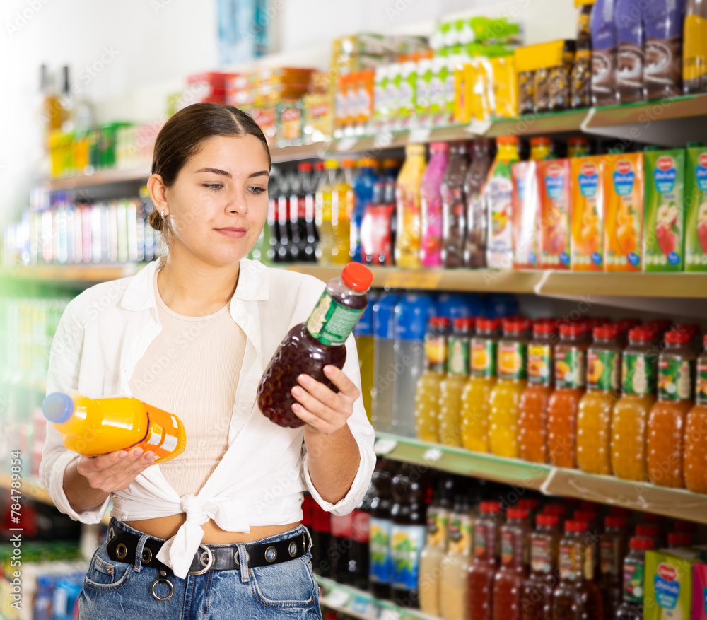 Portrait of young interested girl reading labels on bottles with juices while choosing for soft drinks in grocery store