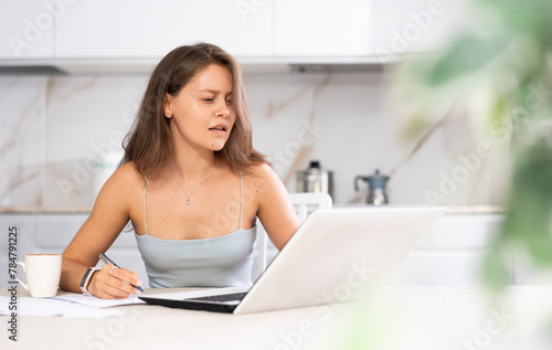 Young woman working remotely using laptop while sitting at table in kitchen