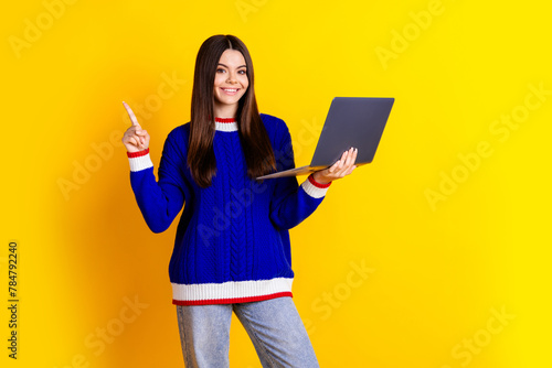 Photo portrait of pretty teen girl hold netbook point empty space wear trendy knitwear blue outfit isolated on yellow color background