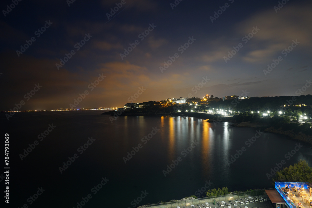 Nighttime View of a Coastal Town With Illuminated Shoreline and Calm Sea.  The serene night sky casts a gentle glow over a coastal town, its warm lights reflecting on the tranquil waters below.