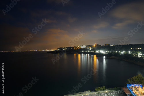 Nighttime View of a Coastal Town With Illuminated Shoreline and Calm Sea. The serene night sky casts a gentle glow over a coastal town, its warm lights reflecting on the tranquil waters below.